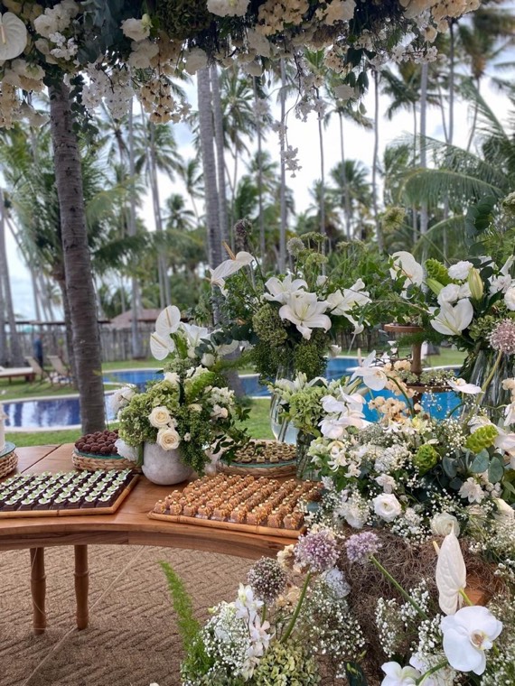 Mesa de doces entre coqueiros com arranjo floral branco