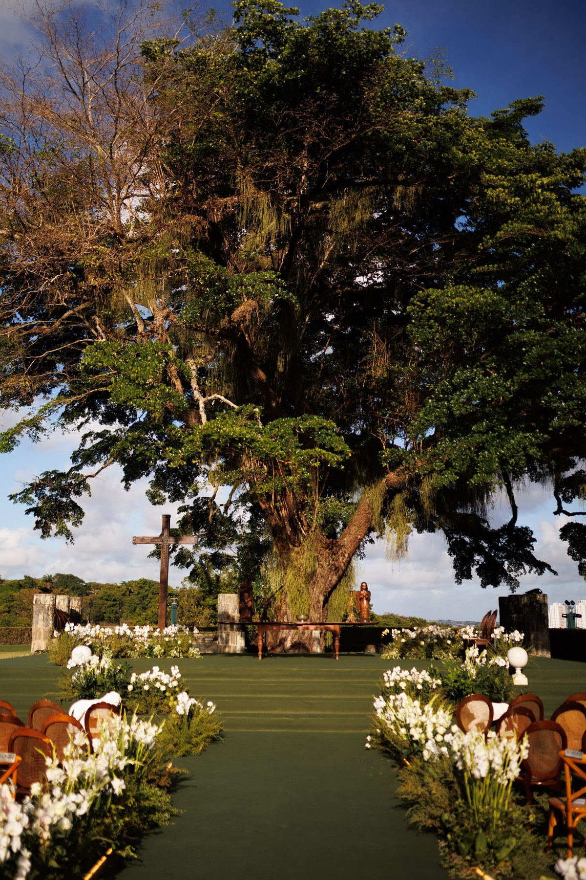 Cerimônia de casamento ao ar livre sob árvore centenária, com corredor florido, cruz rústica e altar cenográfico